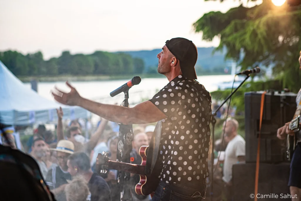 Alex de Basket Case en concert à la Fête de la Musique Alex de Basket Case chante et joue de la guitare en concert à la Fête de la Musique, devant une foule enthousiaste près de l'eau.