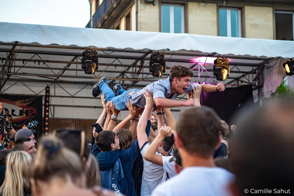 Concert de Basket Case à la fête de la musique avec un public en délire Un jeune homme est porté par la foule lors d'un concert de Basket Case à la fête de la musique, avec la scène en arrière-plan.