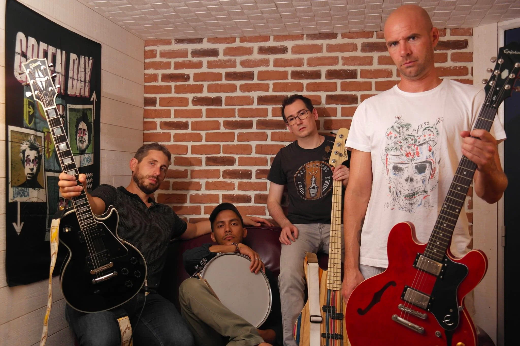 Photo des membres du groupe Basket Case : Cédric à la guitare solo, Amine à la batterie, Damien à la basse, et Alex à la voix et à la guitare rythmique, posant avec leurs instruments devant un mur de briques.