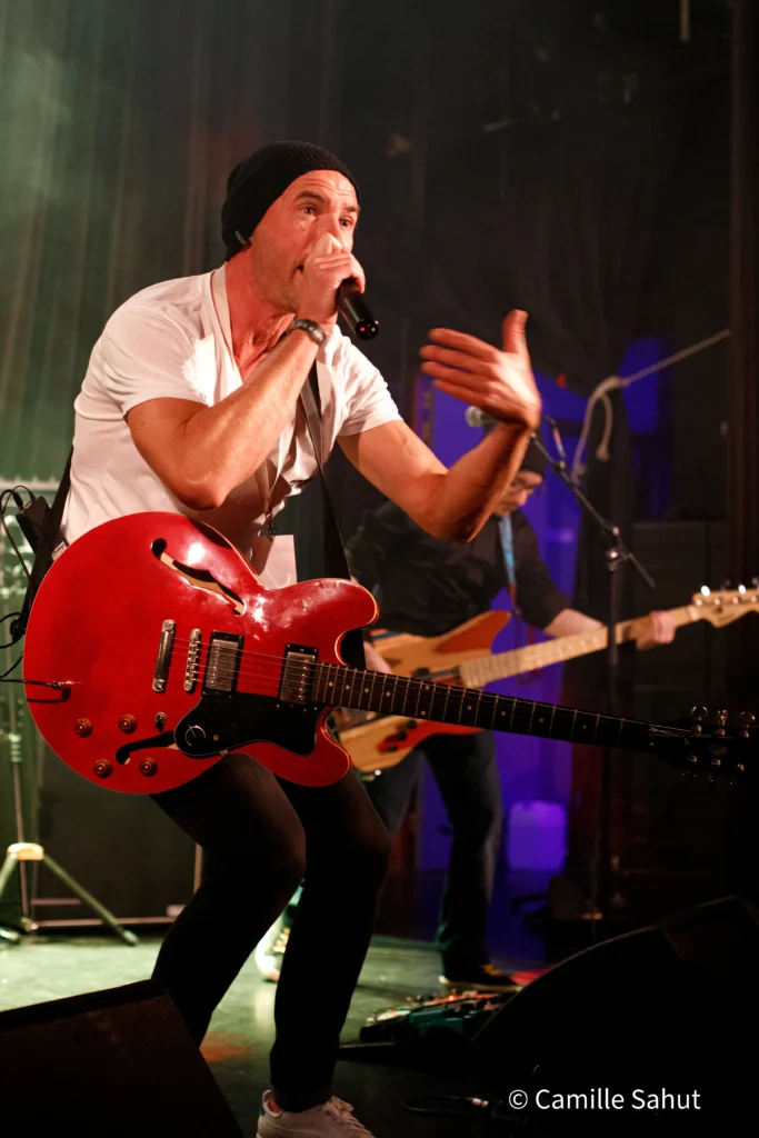 Alex, le chanteur et guitariste du groupe Basket Case, jouant de la guitare rouge sur scène, portant un bonnet noir et une chemise blanche, avec des lumières de scène en arrière-plan.