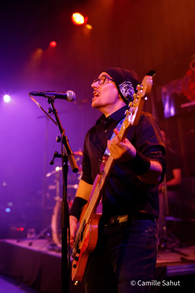 Damien, le bassiste de Basket Case, chantant et jouant de la basse lors d'un concert en live, sous des lumières colorées.